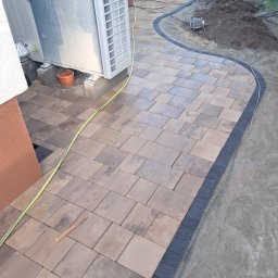 NAWID-NOWOCZESNE BUDOWNICTWO. Brzykcy Napoleon - Zakrzywiona ścieżka z kostki brukowej w odcieniach brązu i szarości, obrzeża z ciemnej kostki, widoczny agregat klimatyzacji na betonowych podstawach, kabel elektryczny oraz narzędzie do układania...