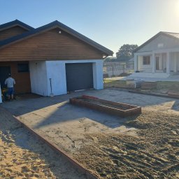 NAWID-NOWOCZESNE BUDOWNICTWO. Brzykcy Napoleon - Przygotowanie podłoża pod kostkę brukową z widocznymi obrzeżami i dwoma budynkami w tle, jeden z garażem, w słoneczny dzień.