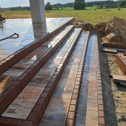 NAWID-NOWOCZESNE BUDOWNICTWO. Brzykcy Napoleon - Kamienne schody zewnętrzne z brązowymi obrzeżami, częściowo mokre, prowadzące do budynku, z widokiem na pole w tle.