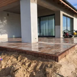 Nowo ułożony taras z brązowej kostki brukowej przed nowoczesnym domem z przesuwnymi drzwiami, widoczna zagęszczarka w tle.