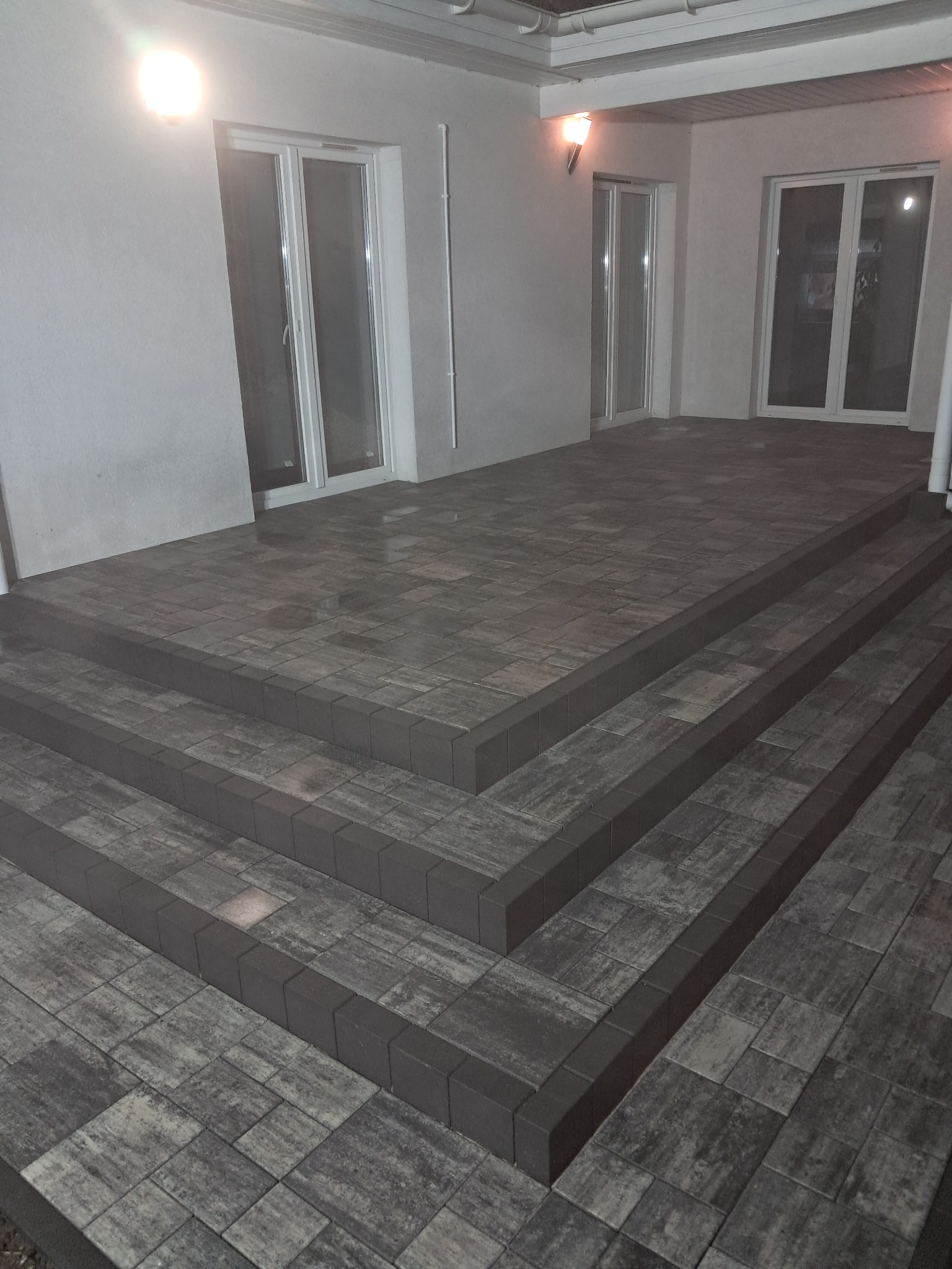 Schody zewnętrzne z kostki brukowej w odcieniach szarości, prowadzące do wejścia budynku z trzema parami drzwi balkonowych, oświetlone lampami zewnętrznymi.