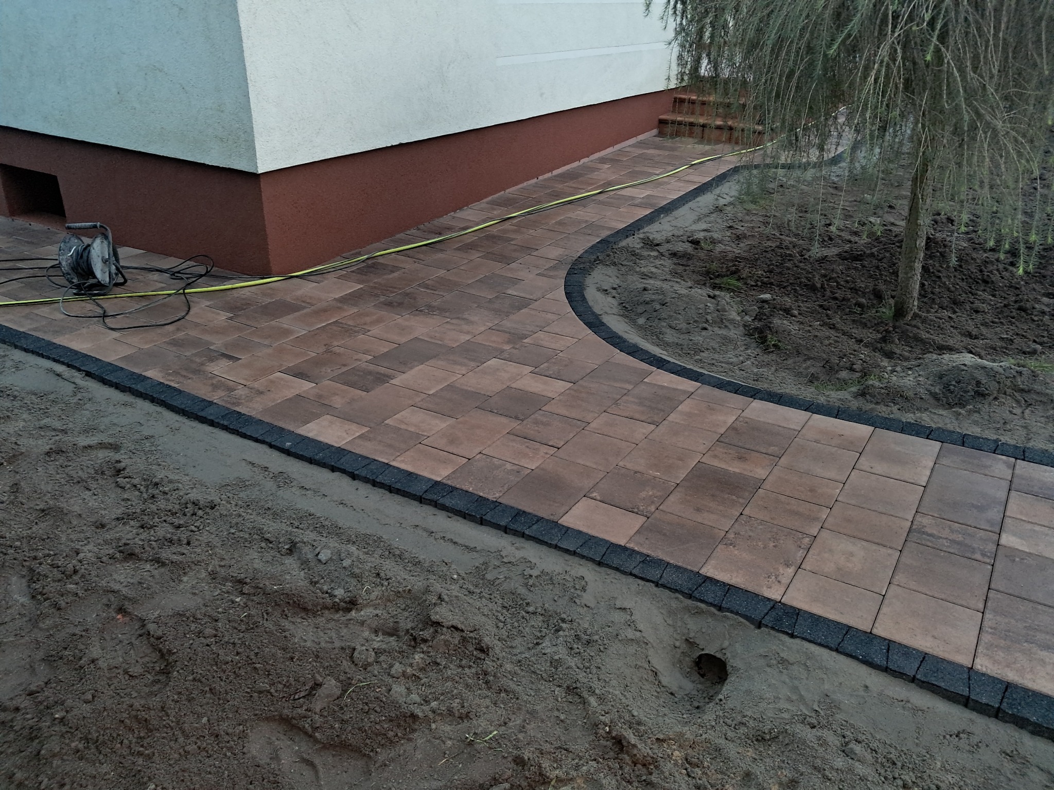 Ścieżka z brązowej kostki brukowej z czarnym obramowaniem, wijąca się obok budynku i drzewa, widoczny zwinięty przedłużacz elektryczny.