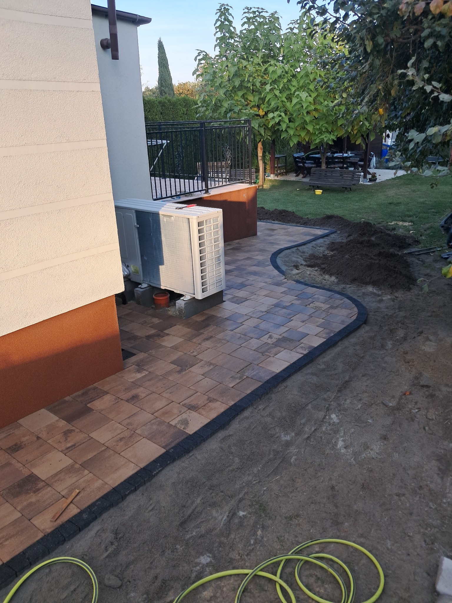 Brązowa kostka brukowa ułożona wzdłuż ściany budynku, z czarnym obrzeżem, prowadząca do trawnika z drzewami i meblami ogrodowymi; widoczny agregat klimatyzacji na zewnątrz budynku.
