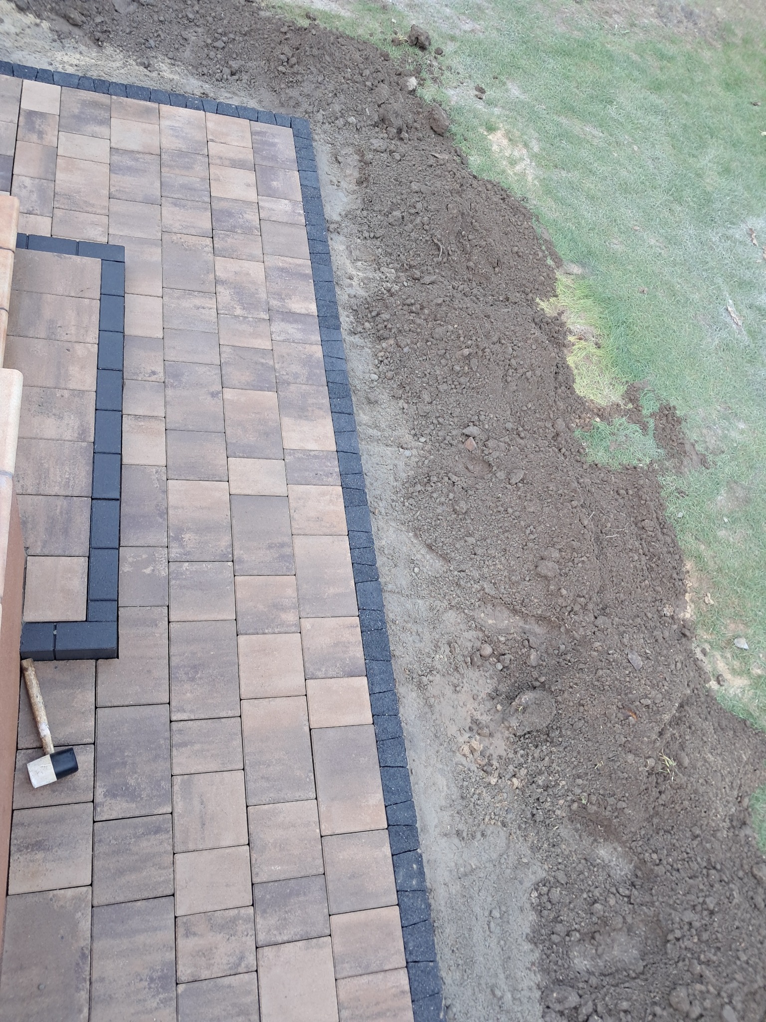 Fragment nowo ułożonej nawierzchni z kostki brukowej w odcieniach brązu, obramowanej ciemnoszarą kostką, widoczny gumowy młotek na skraju oraz świeżo wykopana ziemia i trawa w tle.