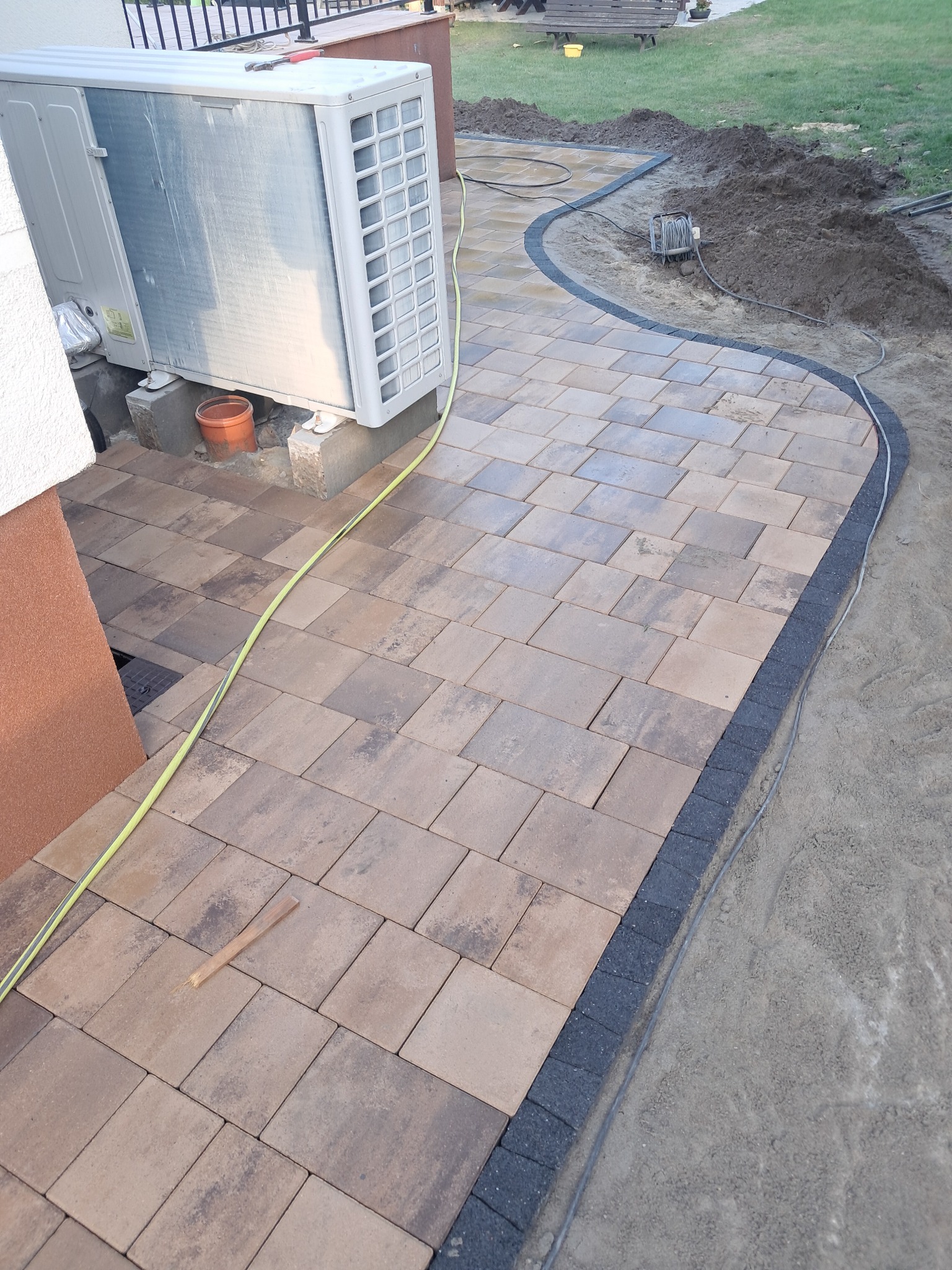 Zakrzywiona ścieżka z kostki brukowej w odcieniach brązu i szarości, obrzeża z ciemnej kostki, widoczny agregat klimatyzacji na betonowych podstawach, kabel elektryczny oraz narzędzie do układania...