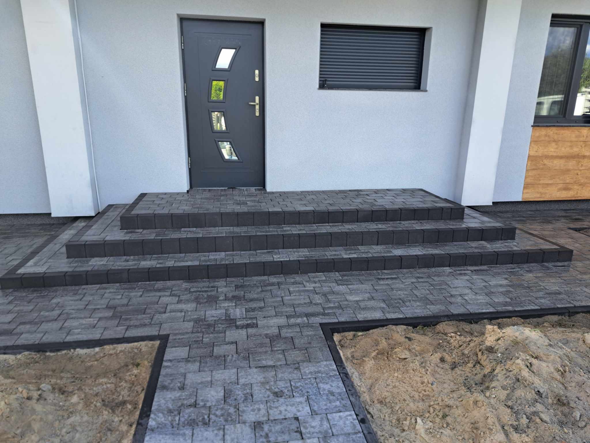 Nowoczesne wejście do domu z szarej kostki brukowej, trzy stopnie prowadzące do ciemnoszarych drzwi z szybkami, obok okno z roletą zewnętrzną, teren wokół częściowo wyłożony kostką, częściowo...
