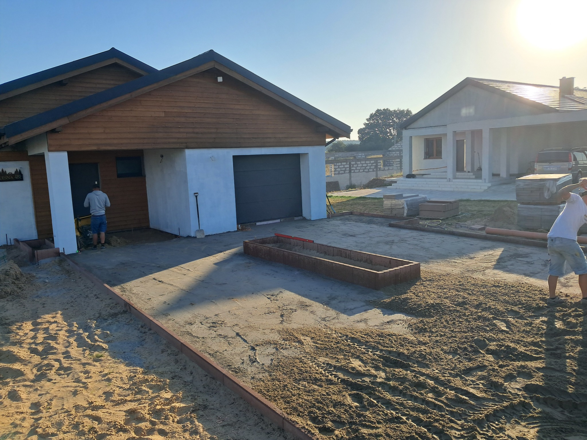 Przygotowanie podłoża pod kostkę brukową z widocznymi obrzeżami i dwoma budynkami w tle, jeden z garażem, w słoneczny dzień.