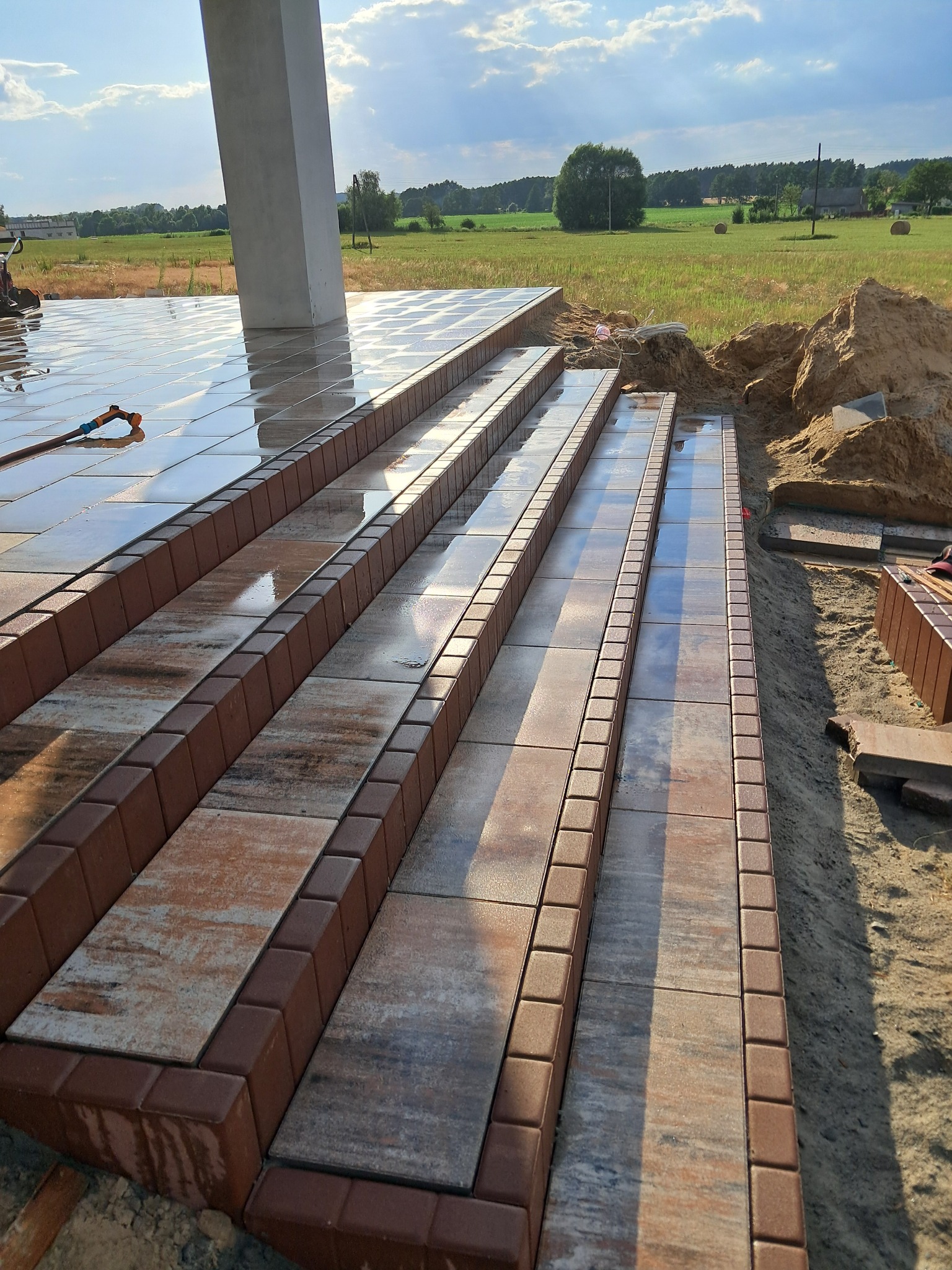 Kamienne schody zewnętrzne z brązowymi obrzeżami, częściowo mokre, prowadzące do budynku, z widokiem na pole w tle.