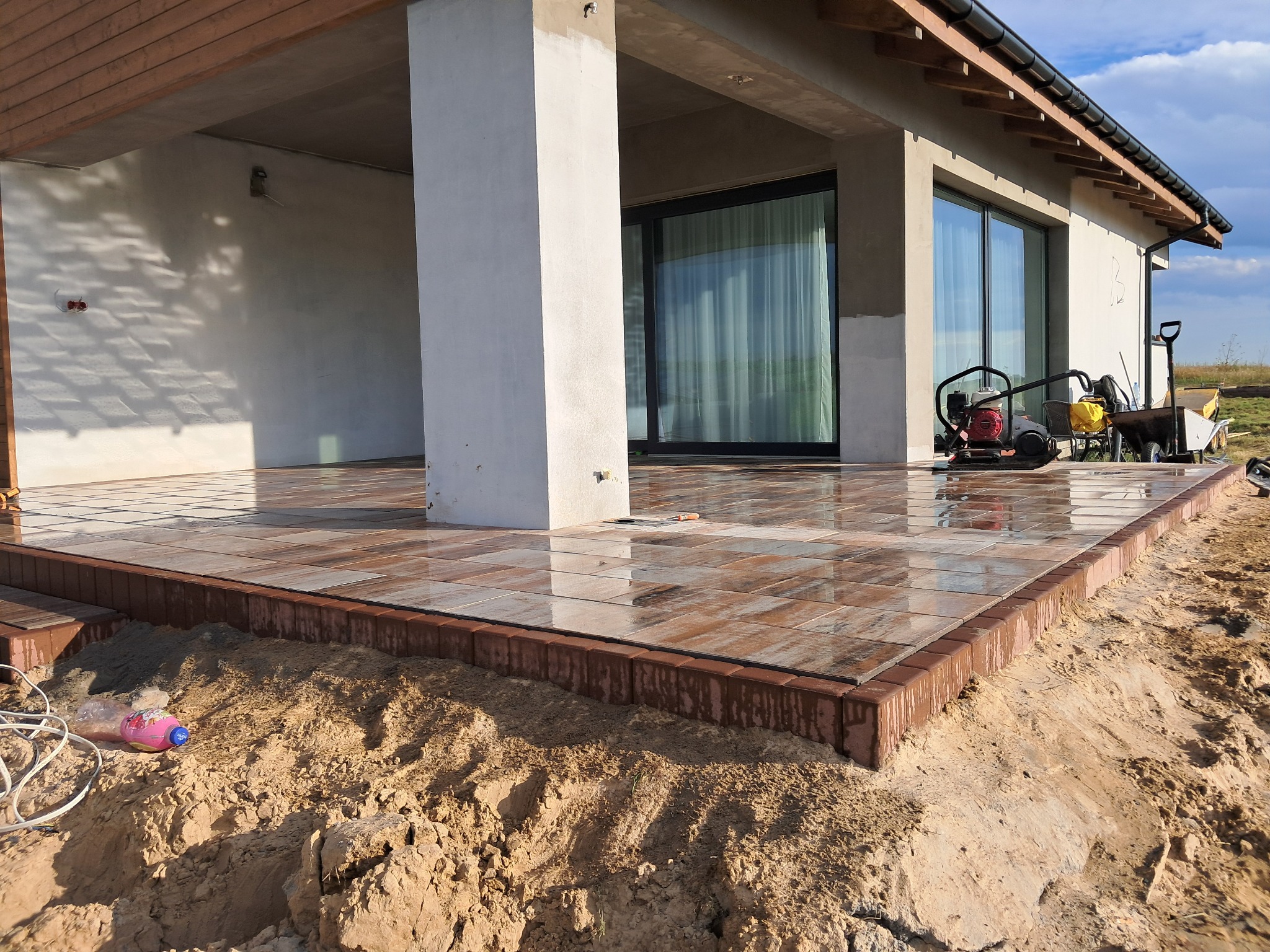 Nowo ułożony taras z brązowej kostki brukowej przed nowoczesnym domem z przesuwnymi drzwiami, widoczna zagęszczarka w tle.