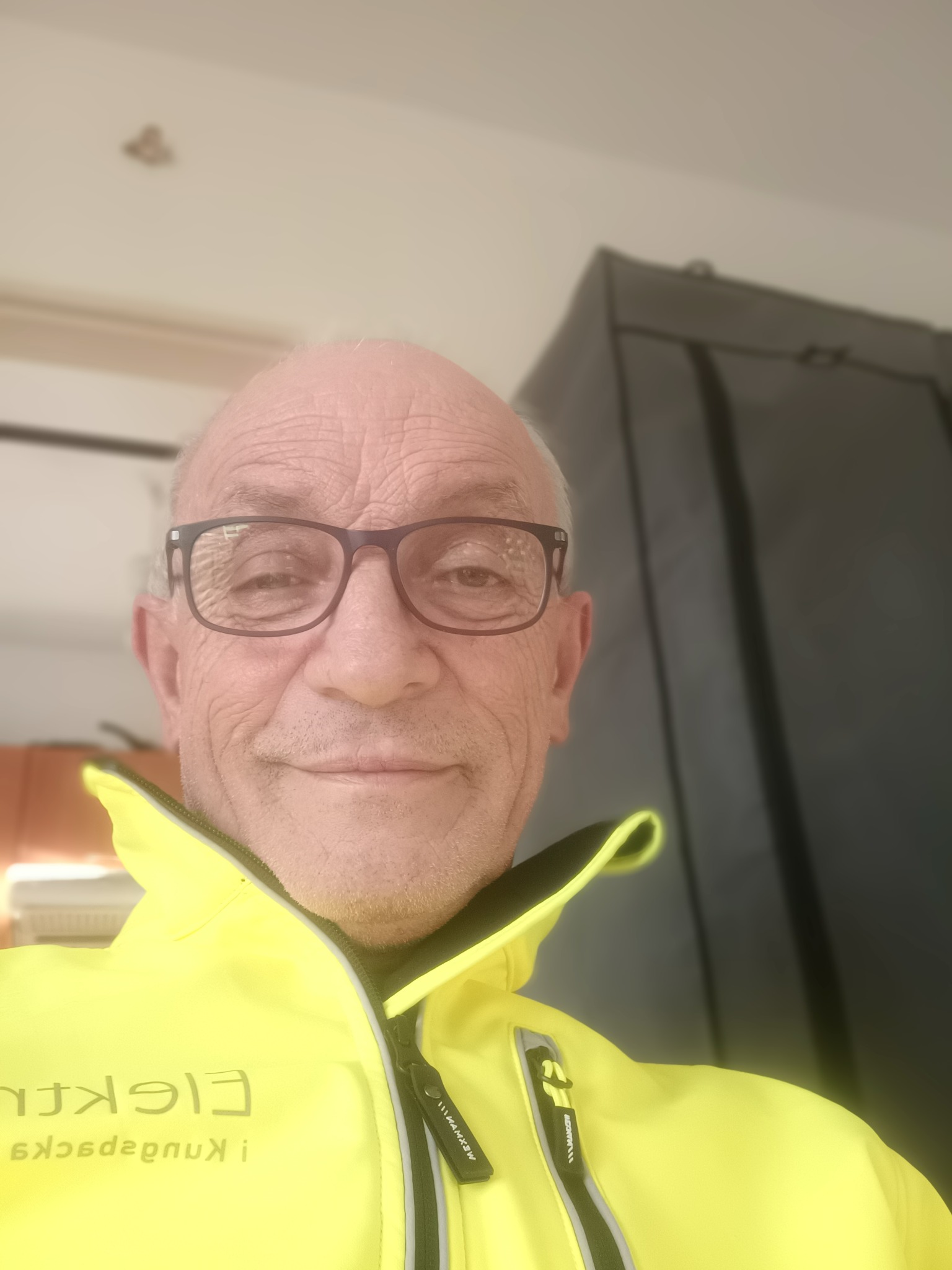 Portret uśmiechniętego elektryka w okularach i jaskrawożółtej kurtce roboczej z widocznym logo firmy.