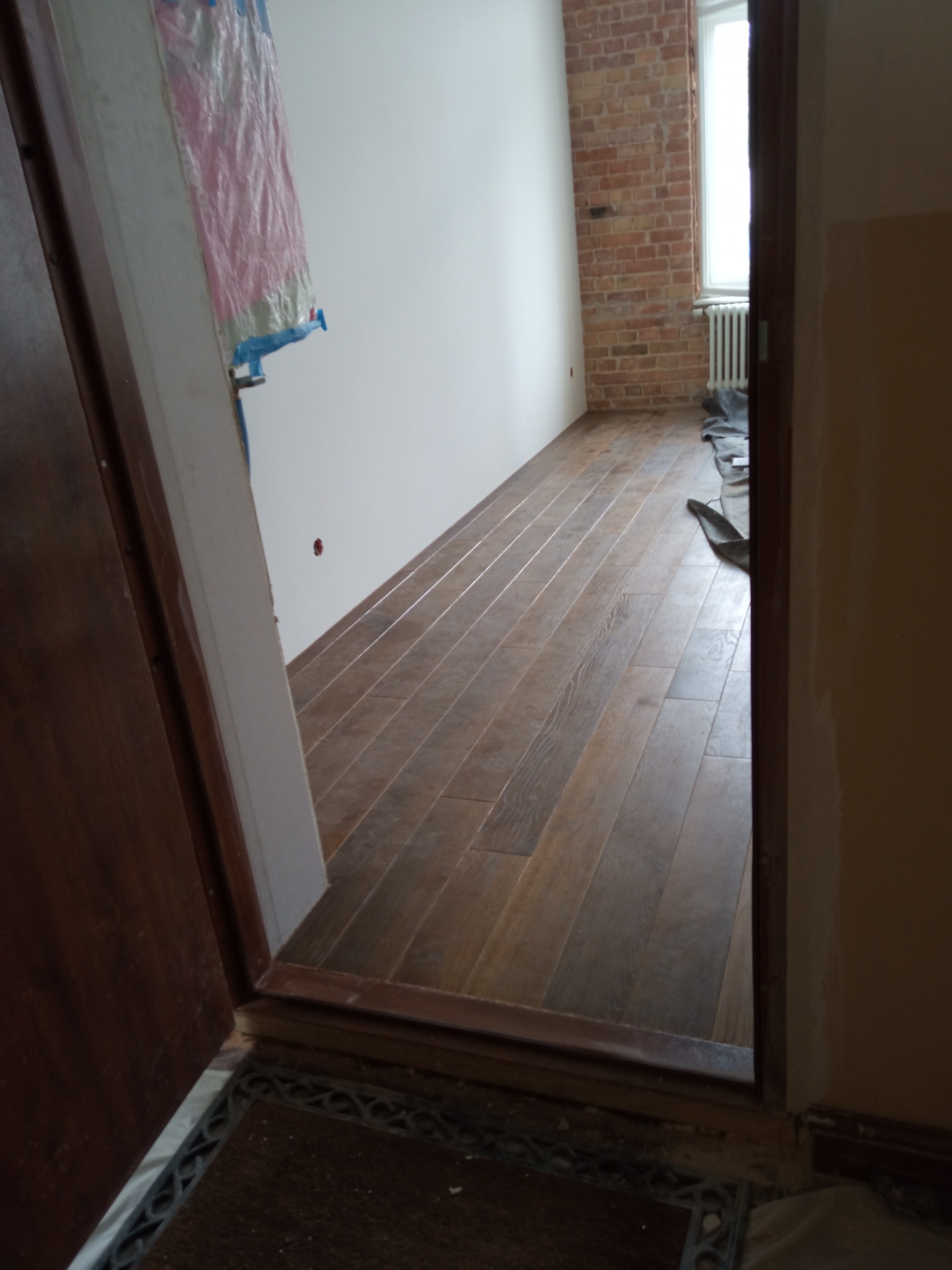 Widok zza otwartych drzwi na świeżo ułożoną, brązową podłogę z paneli imitujących drewno, ściana z białym tynkiem i fragmentem ściany z czerwonej cegły, grzejnik pod oknem.
