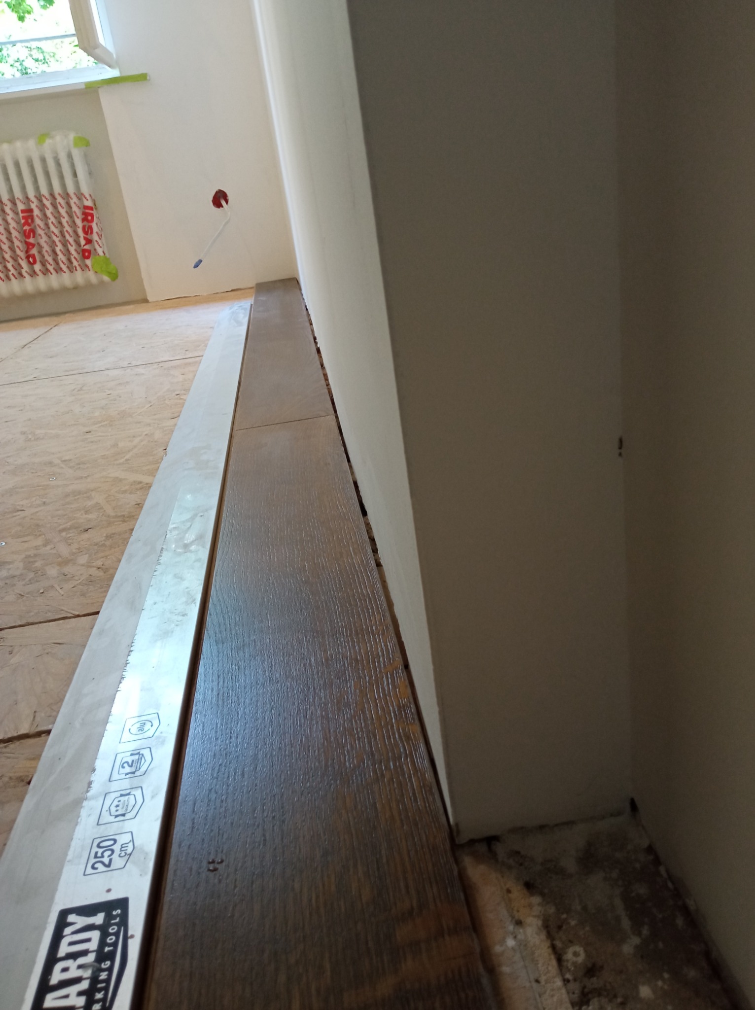 Fragment podłogi z ułożonymi ciemnymi panelami podłogowymi, widoczna listwa przypodłogowa i fragment ściany w trakcie remontu, zabezpieczony grzejnik.