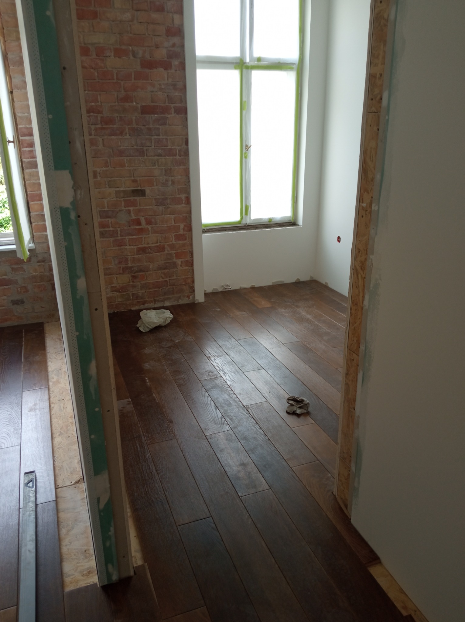 Ciemne, lakierowane deski podłogowe w trakcie układania, widoczne surowe cegły na ścianie i fragmenty konstrukcji ścianki działowej z płyty gipsowo-kartonowej, okna zabezpieczone folią malarską.