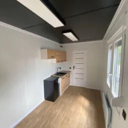 Wnętrze domu modułowego: wykończona kuchnia z drewnianymi szafkami, czarnym blatem i małą czarną lodówką, białe ściany i drzwi, czarny sufit z podłużnymi lampami, podłoga imitująca drewno.