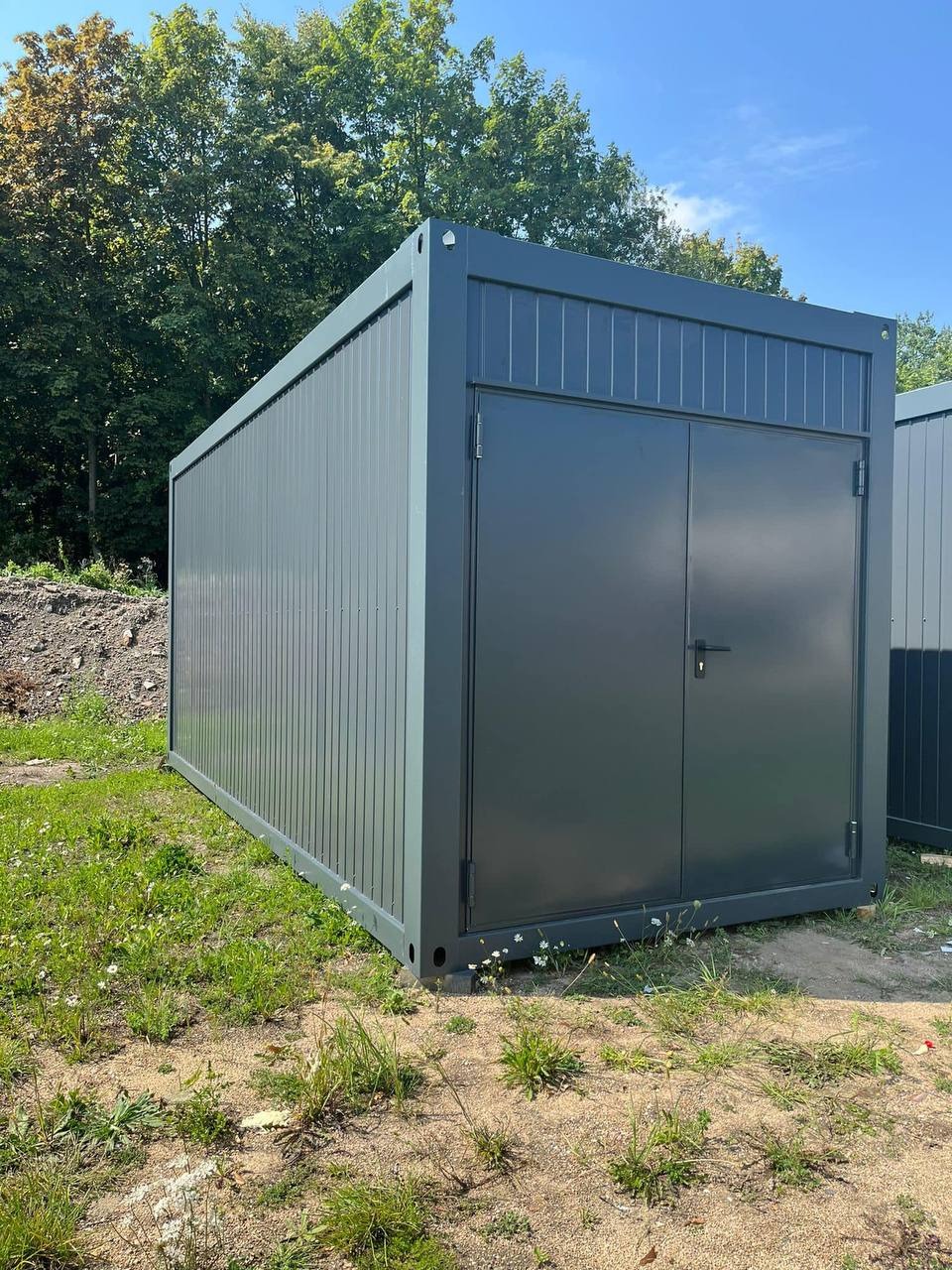Szary kontener mieszkalny z dwuskrzydłowymi drzwiami, ustawiony na trawiastym terenie z widocznym drugim kontenerem w tle.