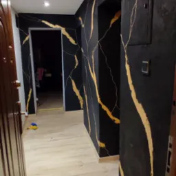 Korytarz z czarnymi ścianami zdobionymi złotymi żyłkami, jasna drewniana podłoga, widoczne drzwi wejściowe i włącznik światła.