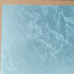 Próbka tynku dekoracyjnego w odcieniach błękitu, prezentowana na tekturze, z widoczną strukturą przypominającą chmury, ułożona na podłodze z paneli drewnopodobnych.