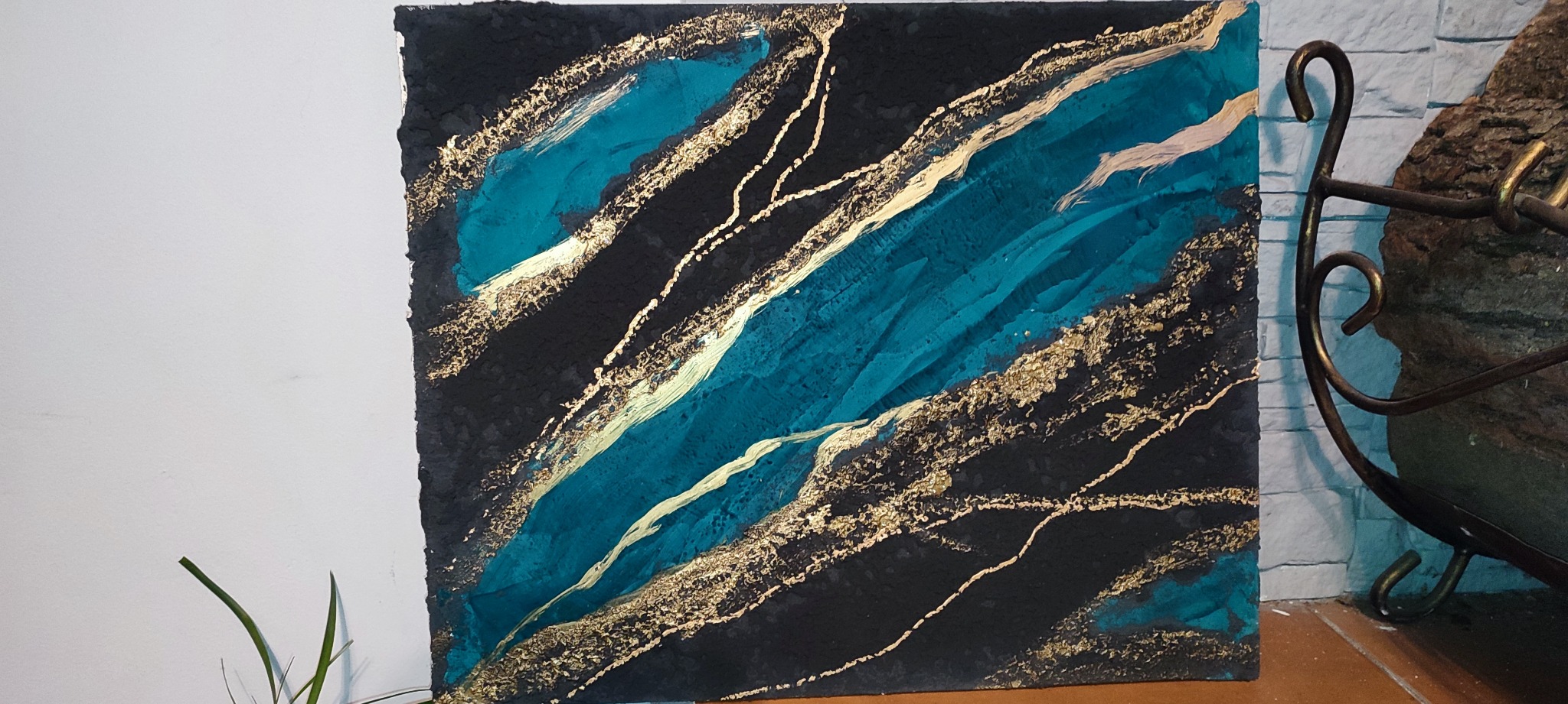 Abstrakcyjna dekoracja ścienna: Turkusowe i złote pasy na czarnym tle z wyraźną fakturą, ustawiona na podłodze obok metalowego stojaka i ściany z cegieł.