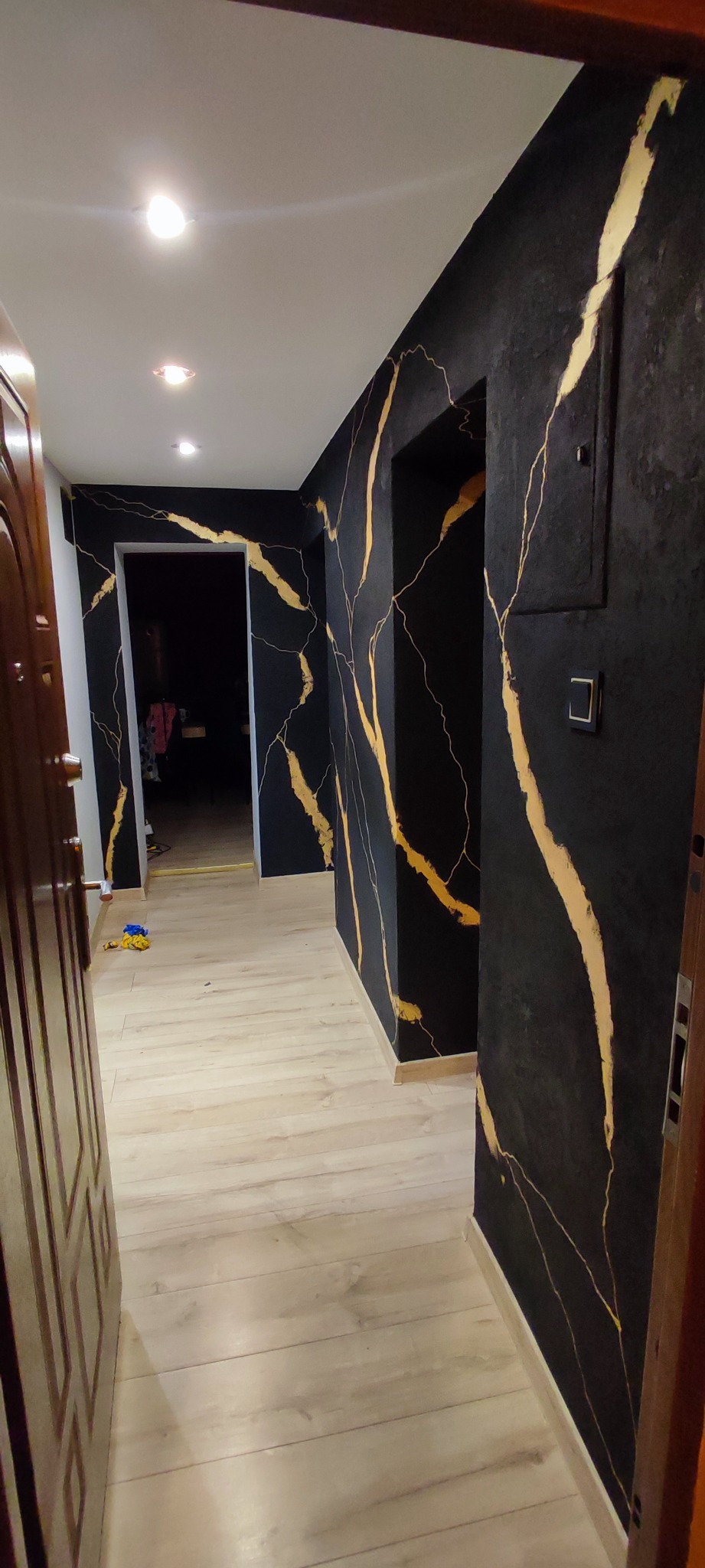 Korytarz z czarnymi ścianami zdobionymi złotymi żyłkami, jasna drewniana podłoga, widoczne drzwi wejściowe i włącznik światła.