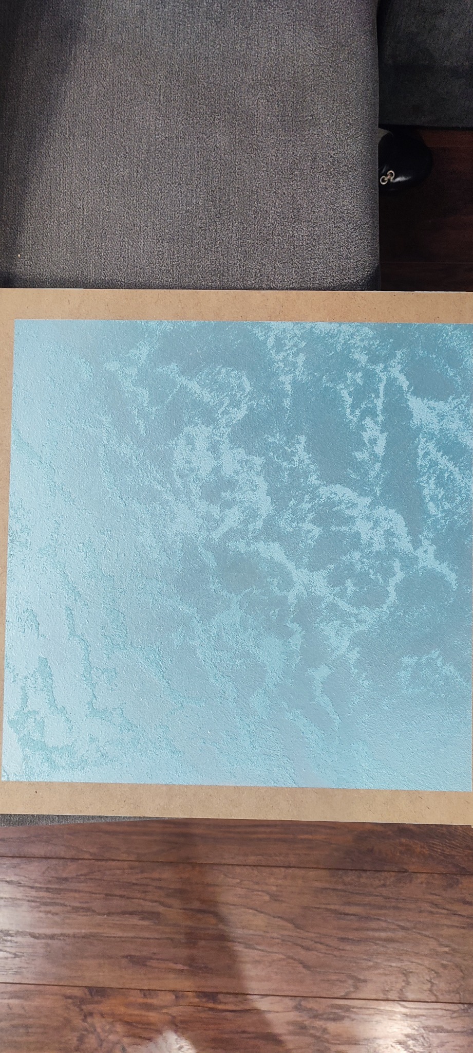 Próbka tynku dekoracyjnego w odcieniach błękitu, prezentowana na tekturze, z widoczną strukturą przypominającą chmury, ułożona na podłodze z paneli drewnopodobnych.