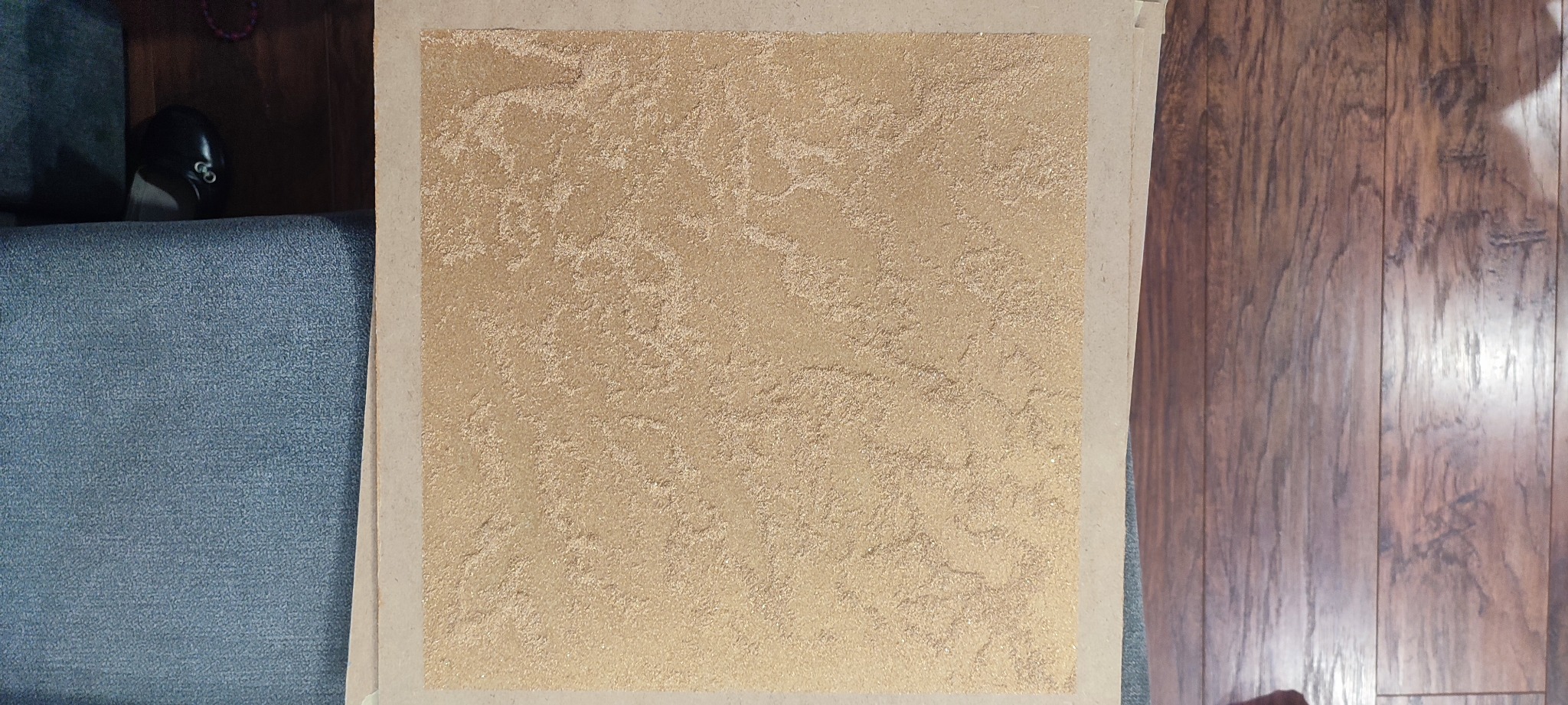 Próbka tynku dekoracyjnego w odcieniach beżu i złota, prezentująca nieregularną, chropowatą fakturę na kartonie, umieszczona obok fragmentu szarej sofy i drewnianej podłogi.