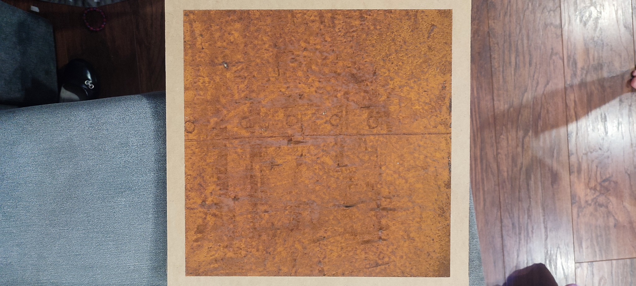 Tekstura imitująca rdzę na płycie z widocznymi odciskami stempli, otoczona beżową ramką, ułożona na podłodze z paneli, obok fragment sofy.