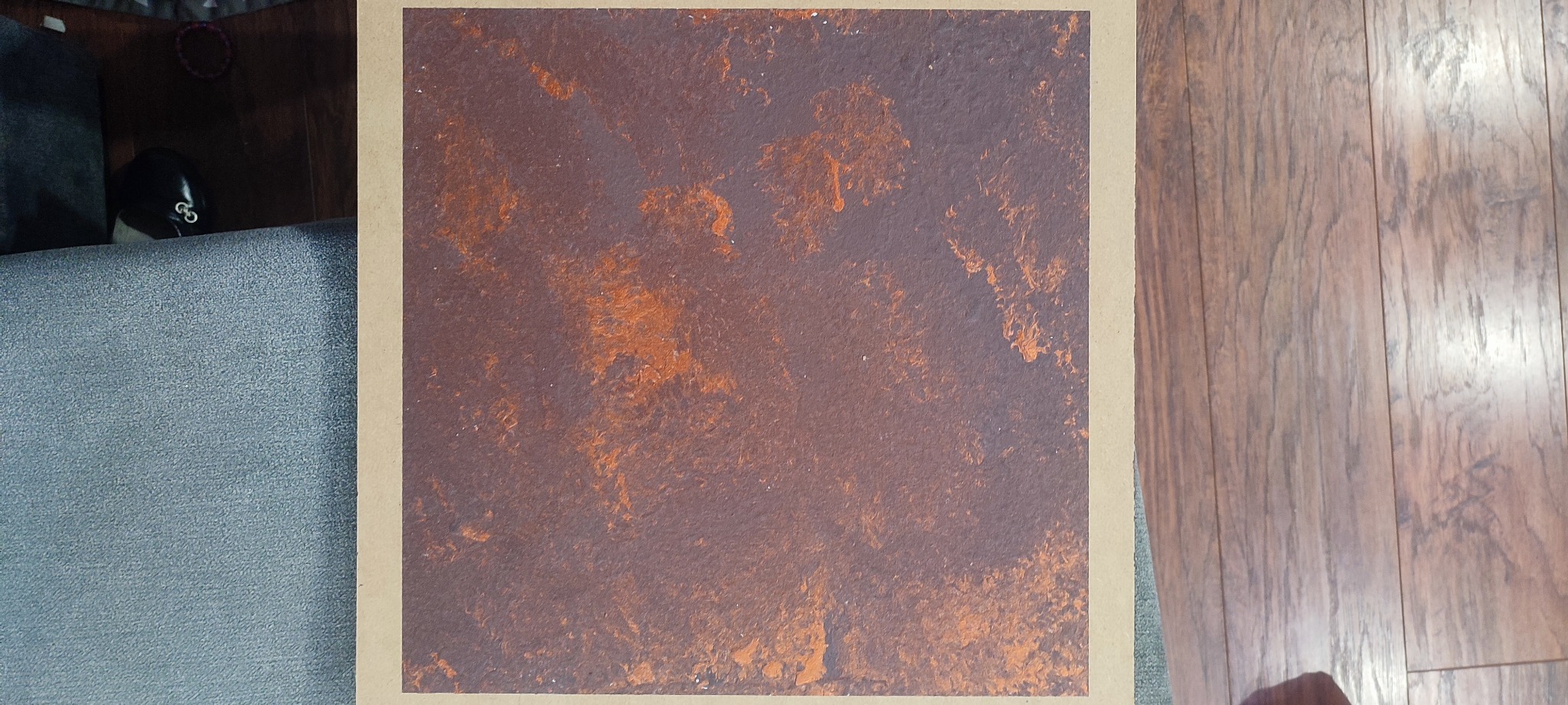 Próbka tynku strukturalnego w odcieniach brązu i rdzy, otoczona beżową ramką, leżąca na podłodze z paneli drewnopodobnych, fragment sofy w tle.