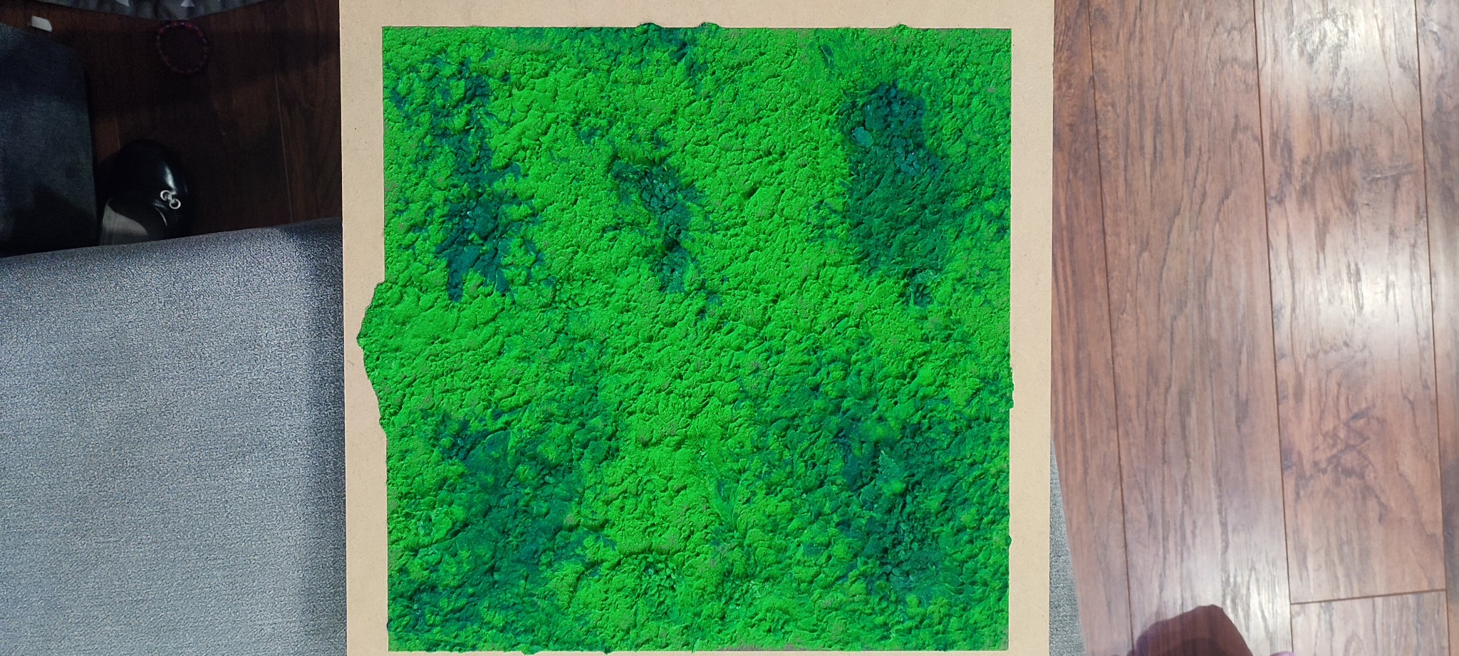Niedokończona, jaskrawozielona tekstura tynku elewacyjnego nałożona na płytę, z widocznymi nierównościami i ciemniejszymi plamami, leżąca na podłodze obok szarej sofy.