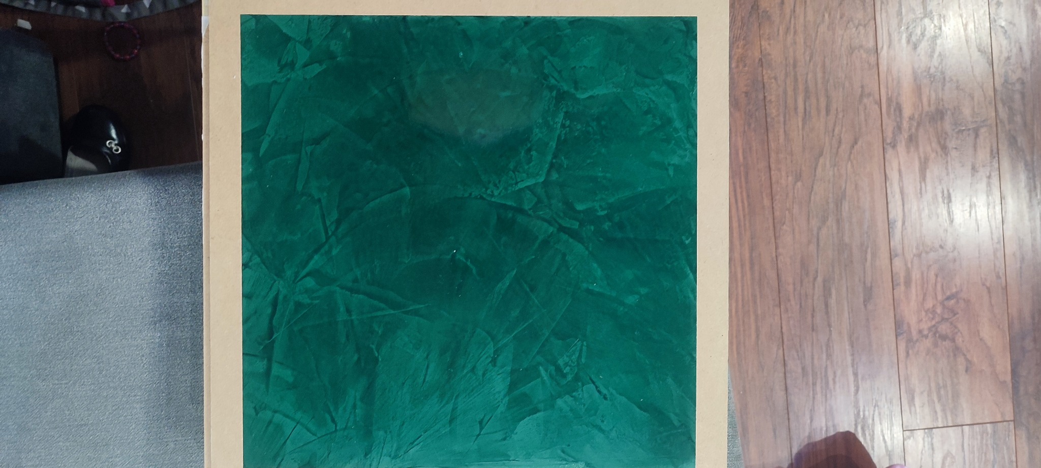 Próbka tynku dekoracyjnego w odcieniach zieleni, prezentowana na kartonowej ramie, z widoczną strukturą imitującą naturalne załamania i nierówności, umieszczona obok drewnianej podłogi.