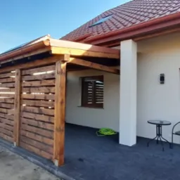 Zadaszony taras z drewnianą ścianą ażurową, przylegający do budynku z czerwonym dachem, widok na ogród w trakcie aranżacji.