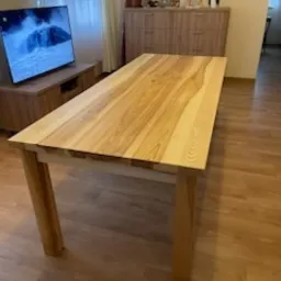 Stół z blatem z jasnego drewna o wyraźnym usłojeniu, ustawiony w salonie z telewizorem i komodą w tle.