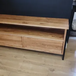 Drewniana szafka RTV z dwoma szufladami i otwartą półką, na czarnych nóżkach, stojąca na podłodze z paneli, obok czarnego stolika i metalowego stojaka na parasole.