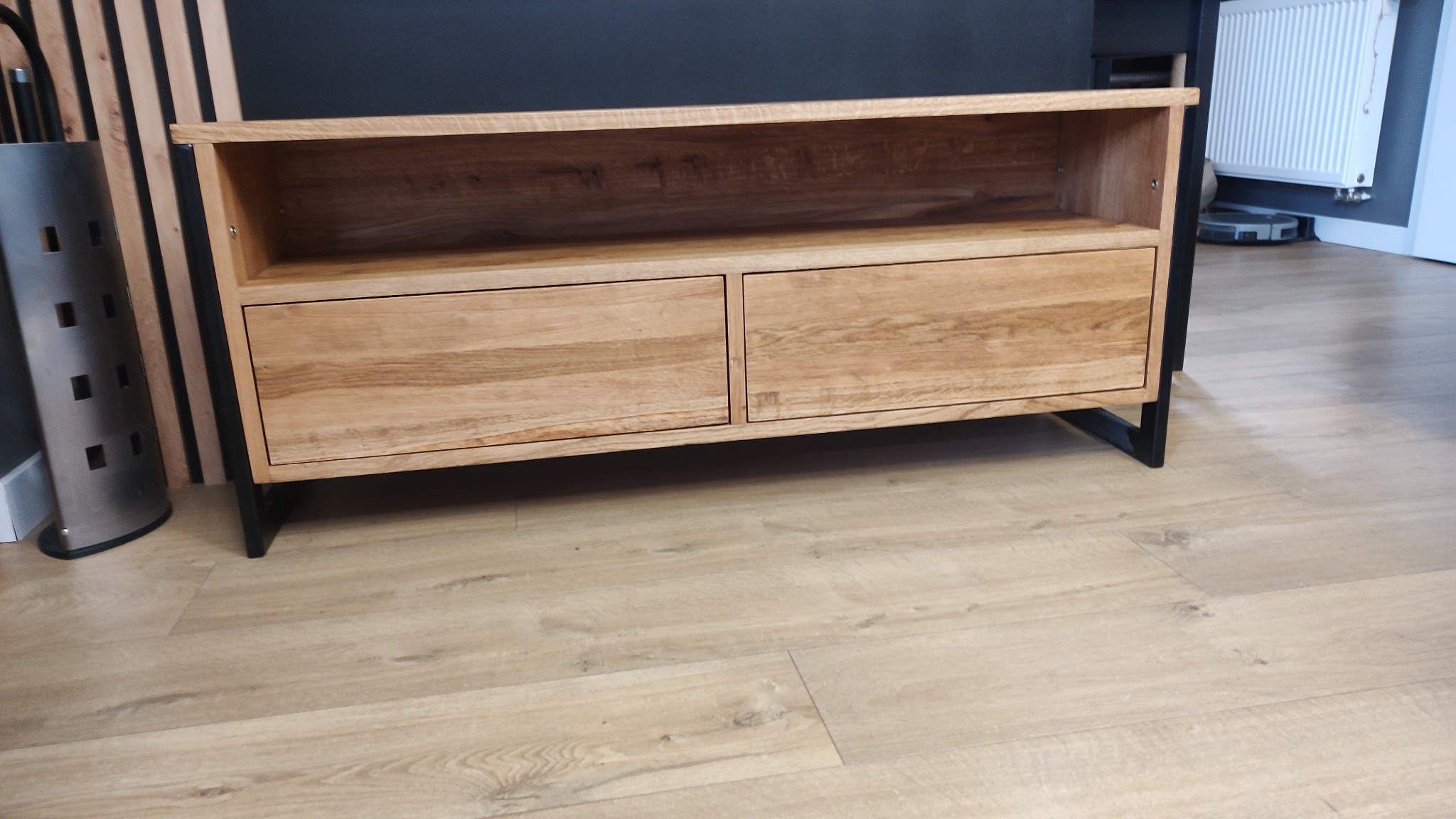 Niska szafka RTV z jasnego drewna dębowego, z dwoma szufladami i otwartą półką, na czarnych metalowych nogach, ustawiona na podłodze z paneli w odcieniach dębu.