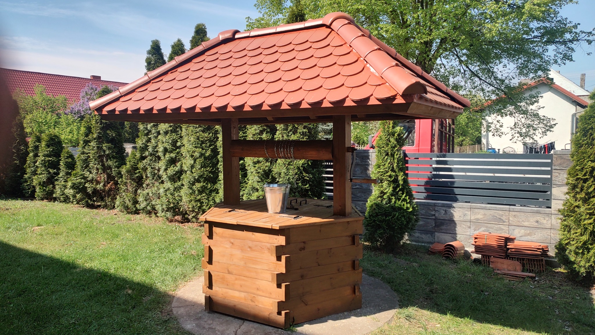 Studnia drewniana zadaszona czerwoną dachówką, wiadro na blacie, otoczona zielonym trawnikiem i tujami, w tle domy.