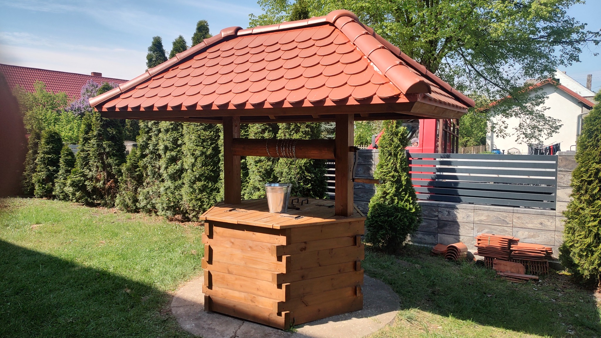 Studnia z drewnianą obudową i czerwoną dachówką, z metalowym wiadrem na blacie, otoczona zielonym trawnikiem i tujami.