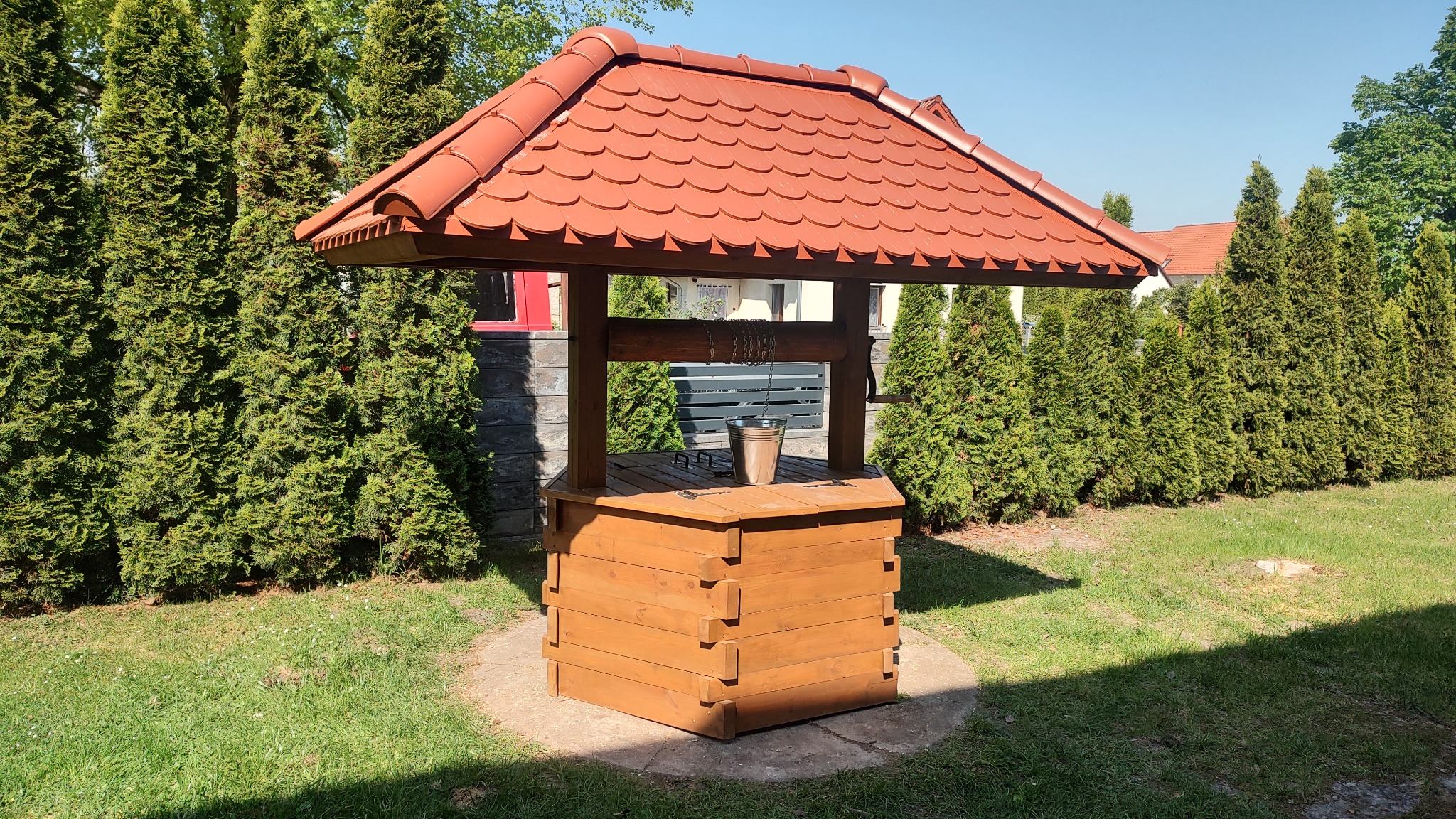 Drewniana studnia ogrodowa z czerwoną dachówką, otoczona tujami na trawniku w słoneczny dzień.