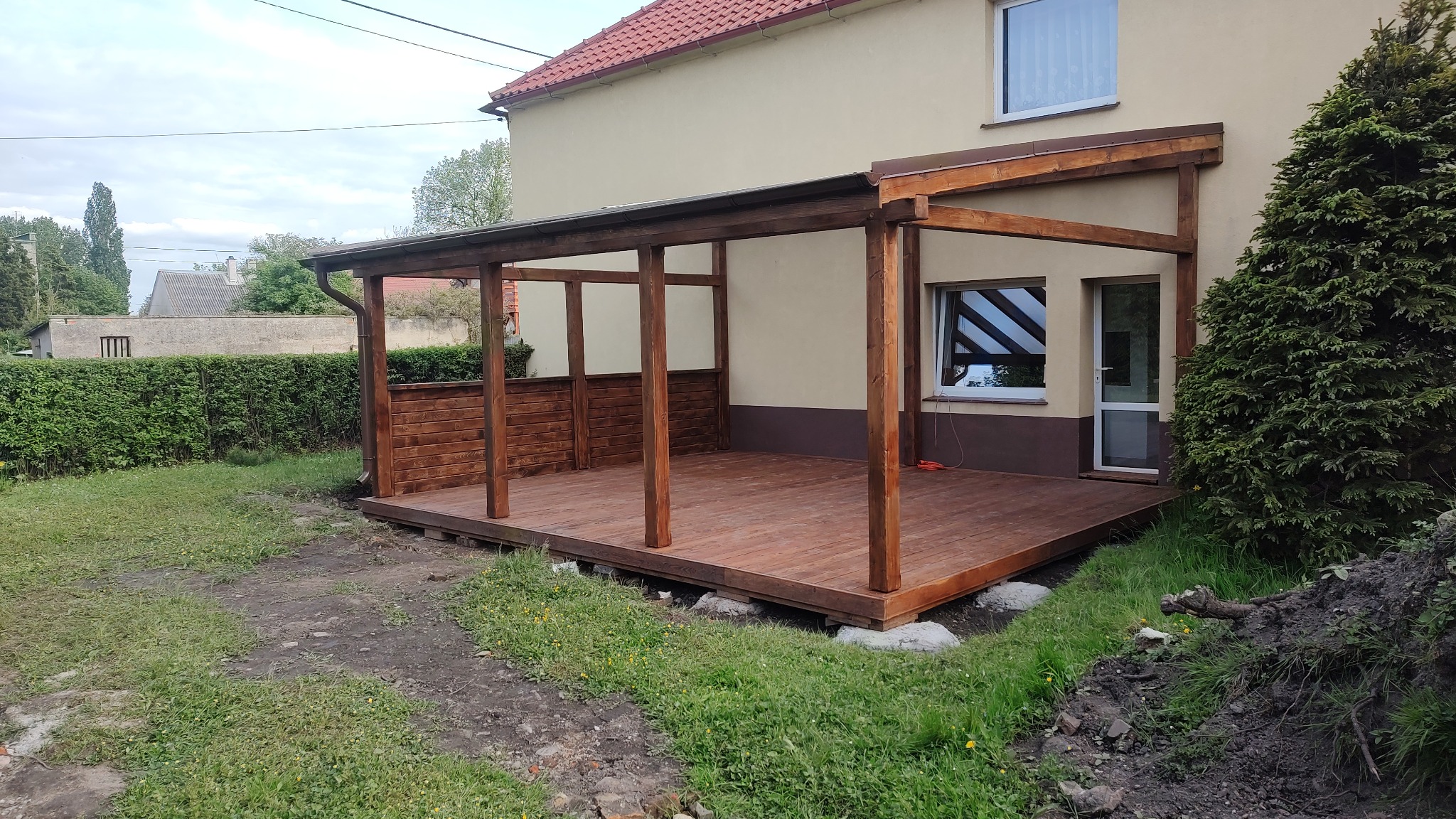 Zadaszony taras drewniany z brązowymi deskami, częściowo obudowany poziomymi panelami, przylegający do budynku z oknem i drzwiami, widok na ogród w trakcie prac ziemnych.
