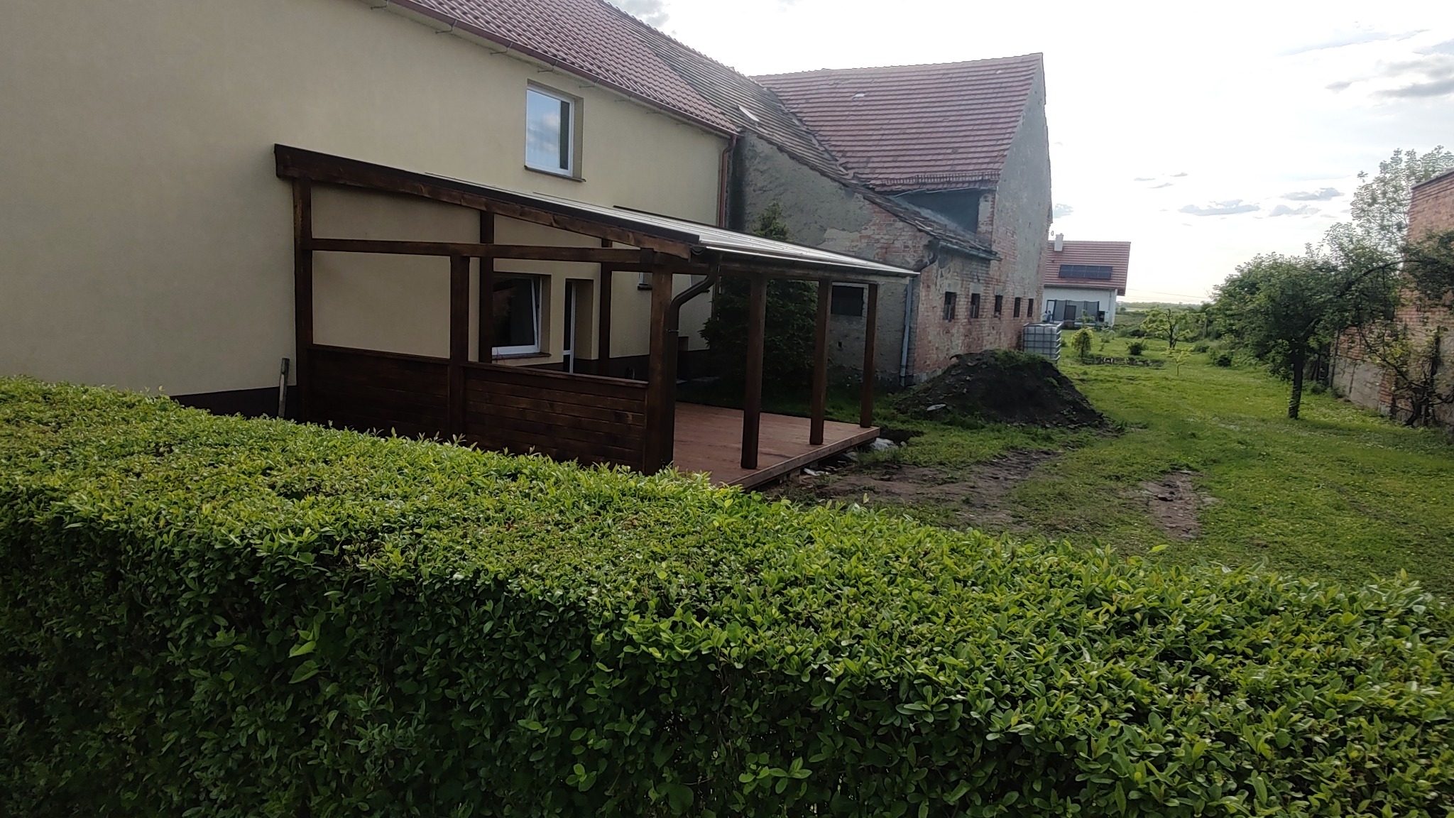 Zadaszony taras z drewnianą podłogą i balustradą, przylegający do budynku mieszkalnego, widoczny zza żywopłotu.