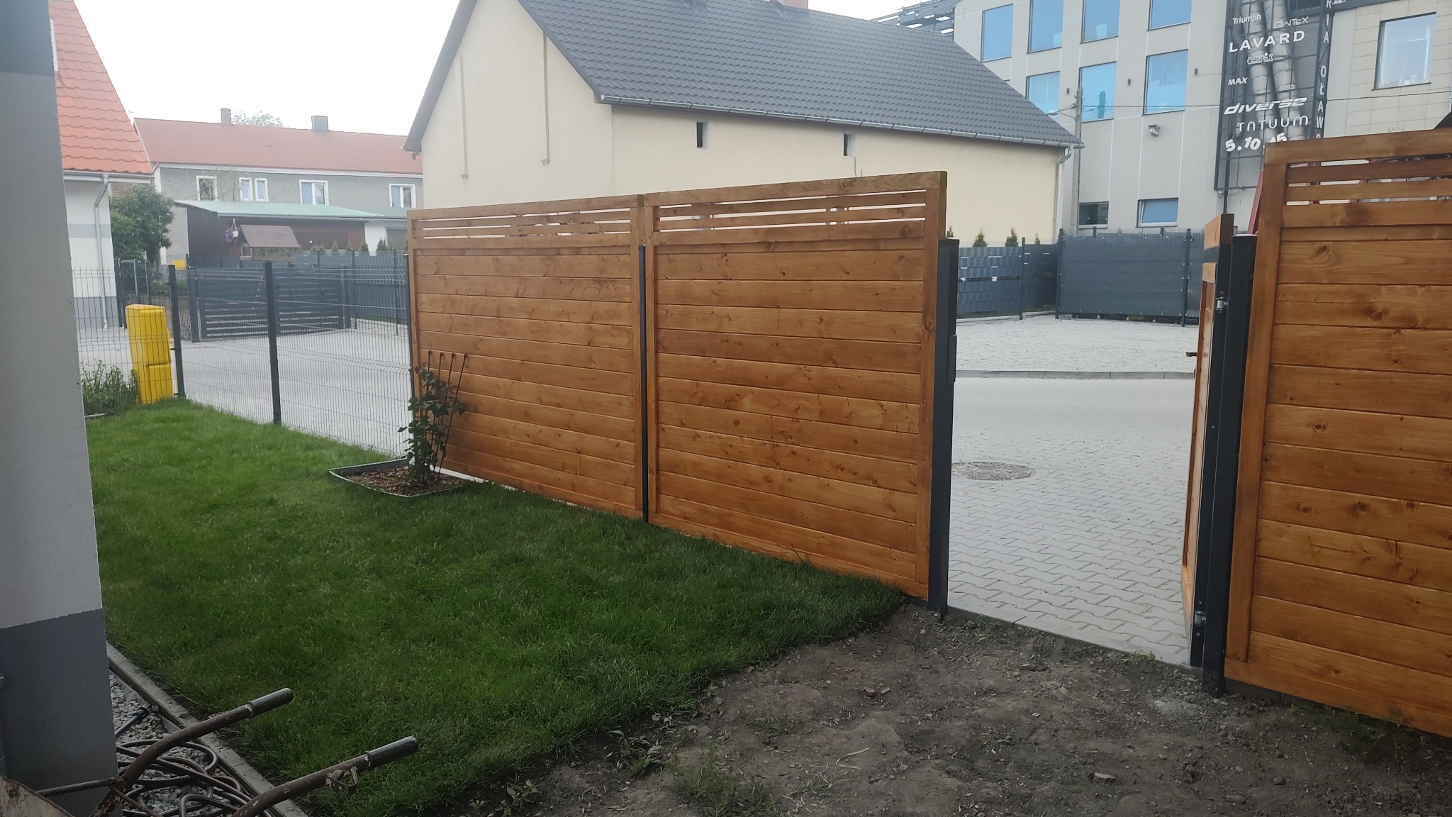 Nowoczesne ogrodzenie z drewnianych paneli, pomalowane na ciepły brąz, z czarnymi metalowymi słupkami, oddzielające trawnik od chodnika z kostki brukowej.