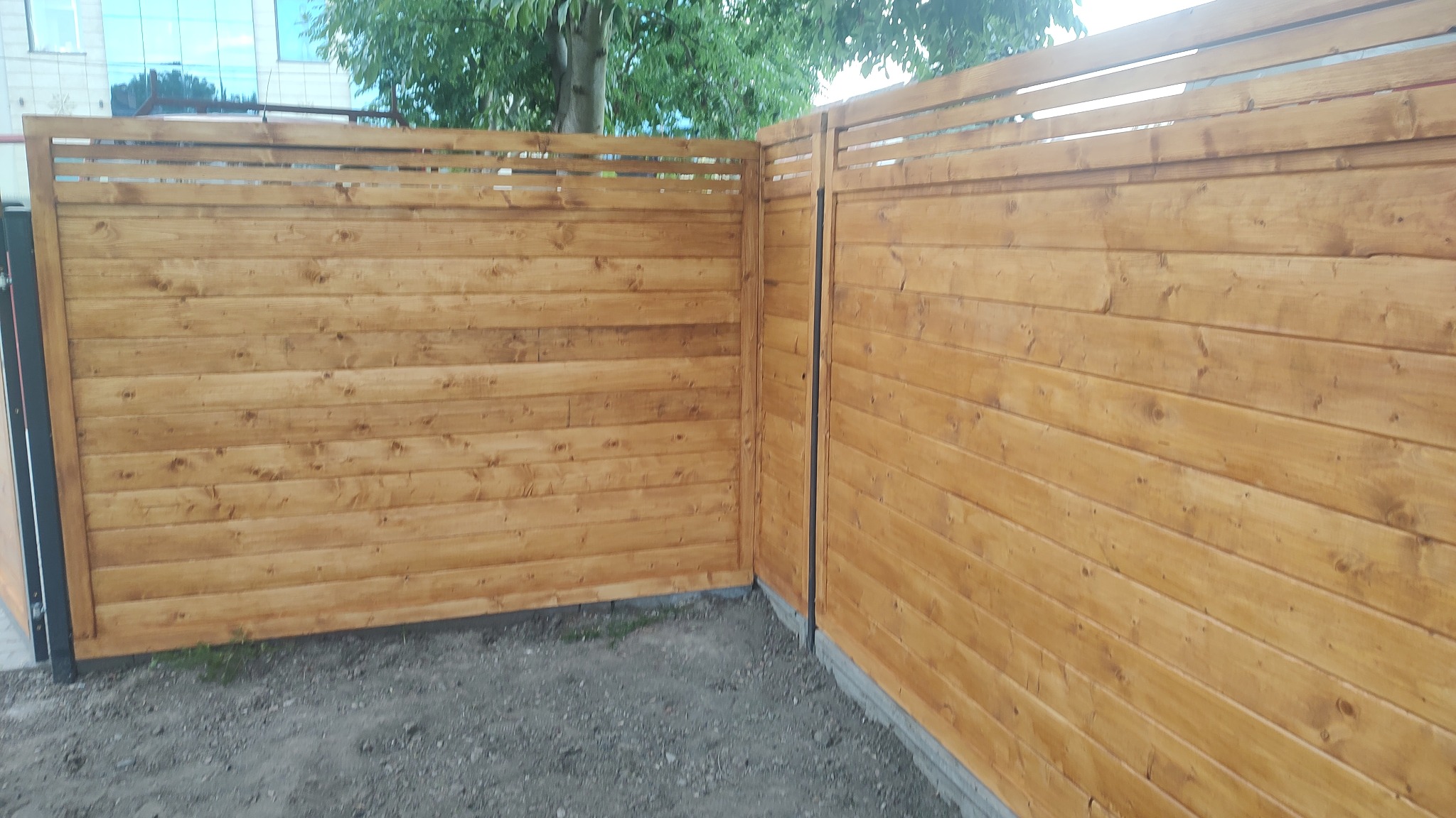 Fragment nowo wybudowanego, poziomego ogrodzenia z impregnowanych desek w odcieniach brązu, z widocznymi słojami, z lekkimi prześwitami między deskami, na tle budynku i drzew