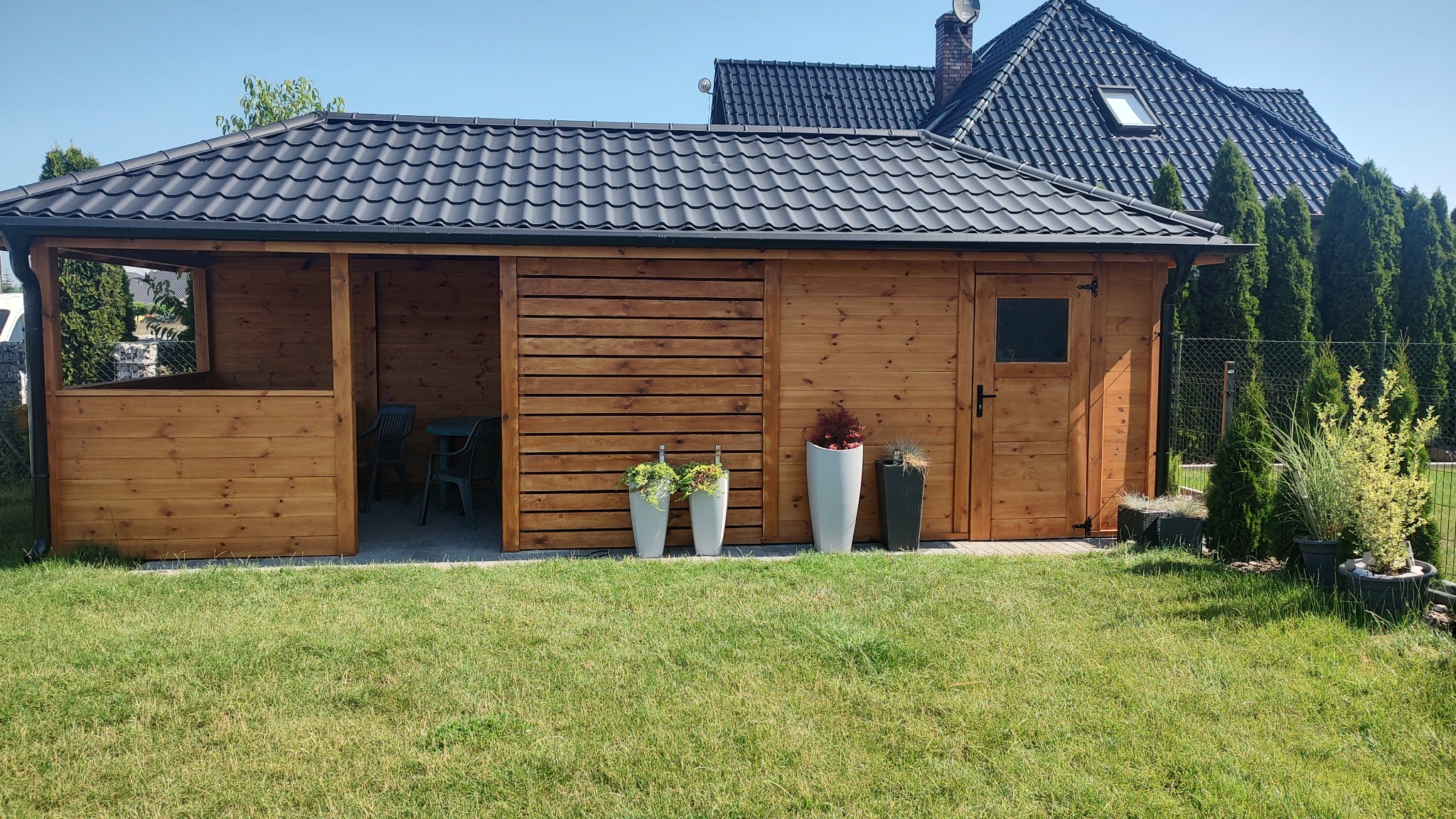 Zadaszona konstrukcja ogrodowa z drewna, z przesuwanym panelem bocznym, czarnym dachem i donicami z roślinami na tle zielonego trawnika i domu.