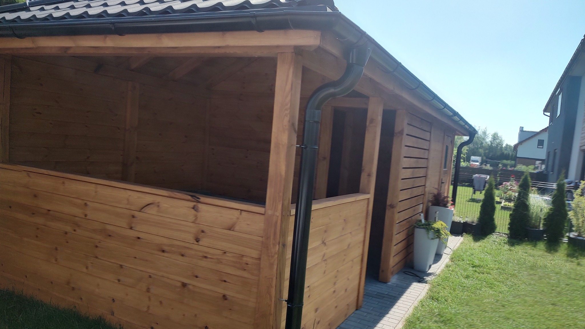 Zadaszona konstrukcja z drewna z czarną rynną, widoczna z zewnątrz na tle zadbanego ogrodu z tujami i budynkami mieszkalnymi.