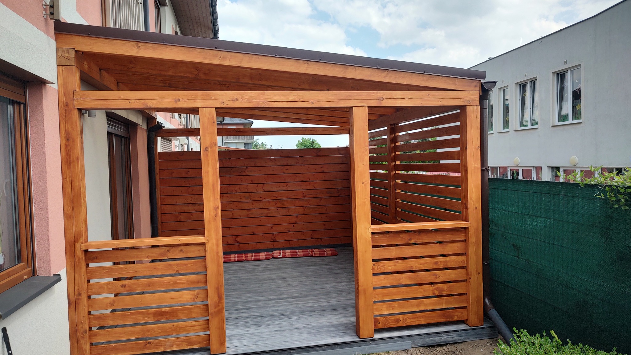 Zadaszona konstrukcja z drewna, przylegająca do budynku, z drewnianymi panelami bocznymi i szarą podłogą kompozytową, widoczne poduszki podłogowe.