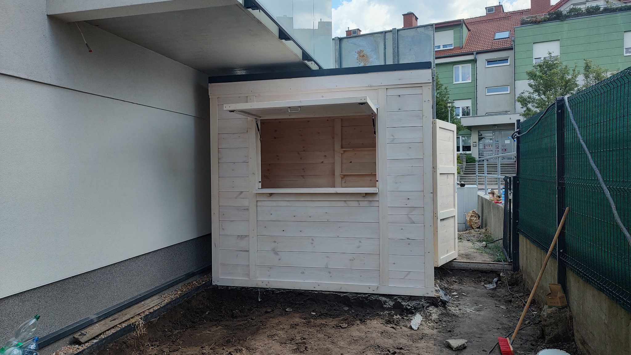 Surowa, jasna drewniana budka z otwieranym okienkiem, ustawiona przy ścianie budynku w trakcie montażu, widoczne narzędzia i niezagospodarowany teren wokół.