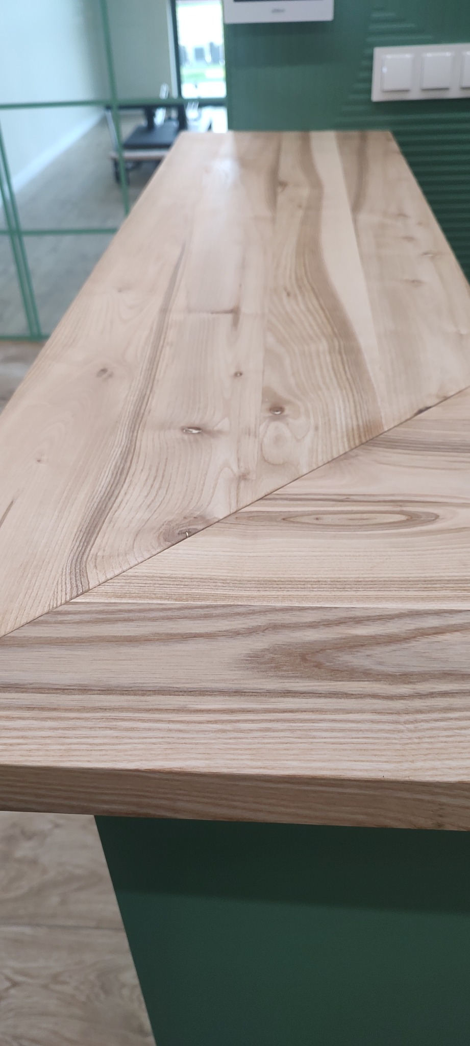 Jasny blat z drewna jesionowego, z wyraźnym usłojeniem, połączony pod kątem, na tle zielonej szafki.