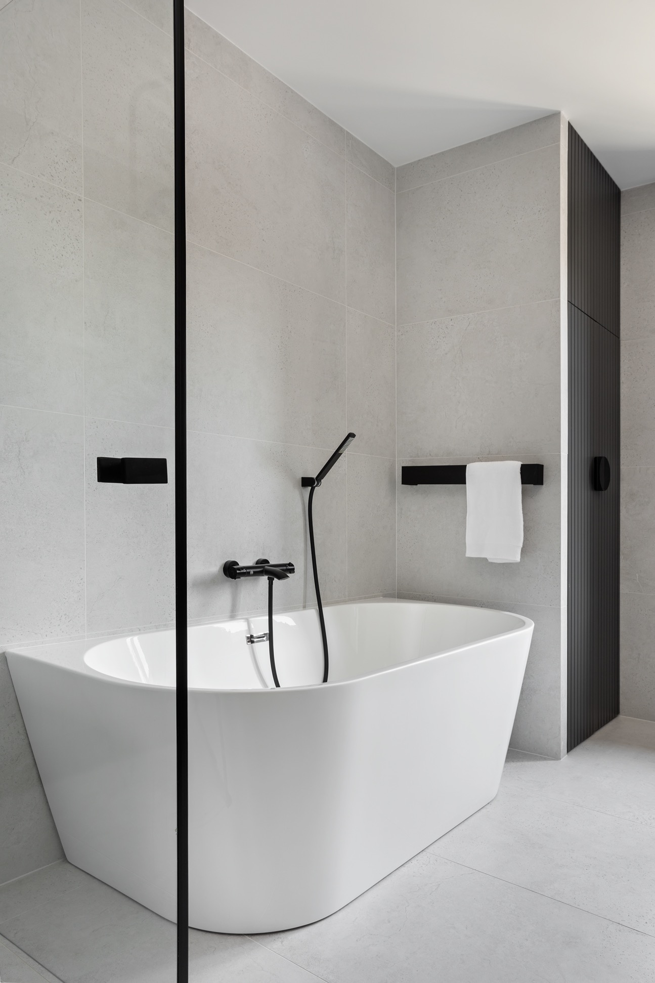 Minimalistyczna łazienka z wolnostojącą białą wanną, czarną armaturą i szklaną ścianką działową, ręcznik na czarnym wieszaku, szare płytki, czarne lamele dekoracyjne.