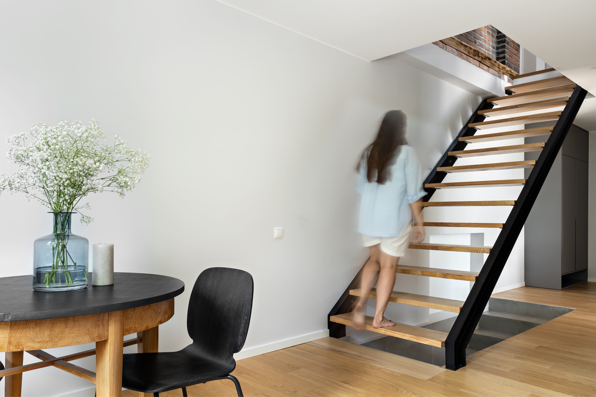 Wnętrze mieszkania z drewnianymi schodami o czarnej konstrukcji, drewnianą podłogą i okrągłym stołem z kwiatami w wazonie oraz rozmyta sylwetka kobiety wchodzącej po schodach.