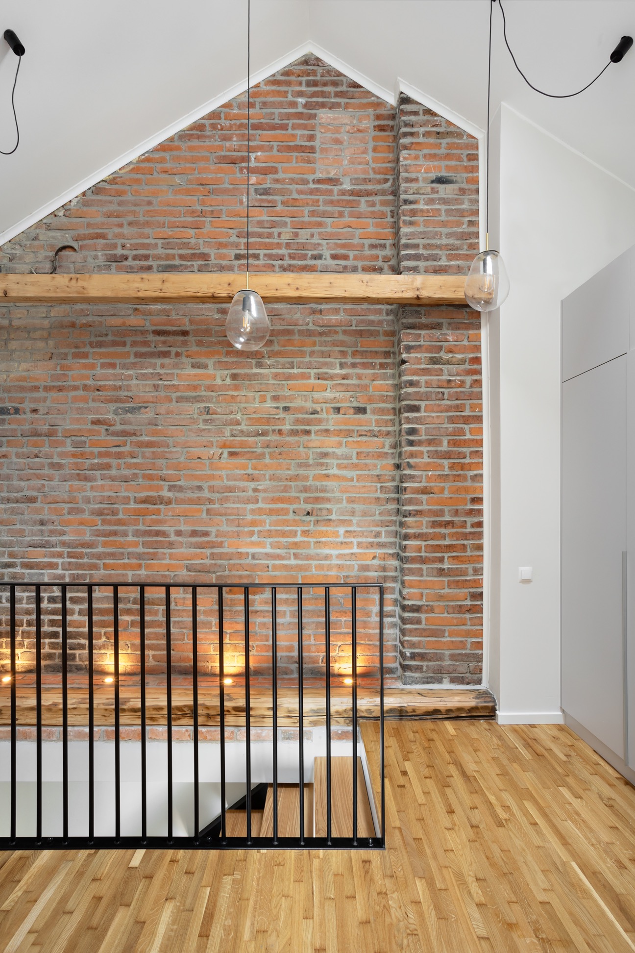 Loftowe wnętrze z ceglaną ścianą, drewnianą podłogą i czarną balustradą. Dwie szklane lampy wiszące oświetlają przestrzeń. Widoczne schody z drewnianymi stopniami.
