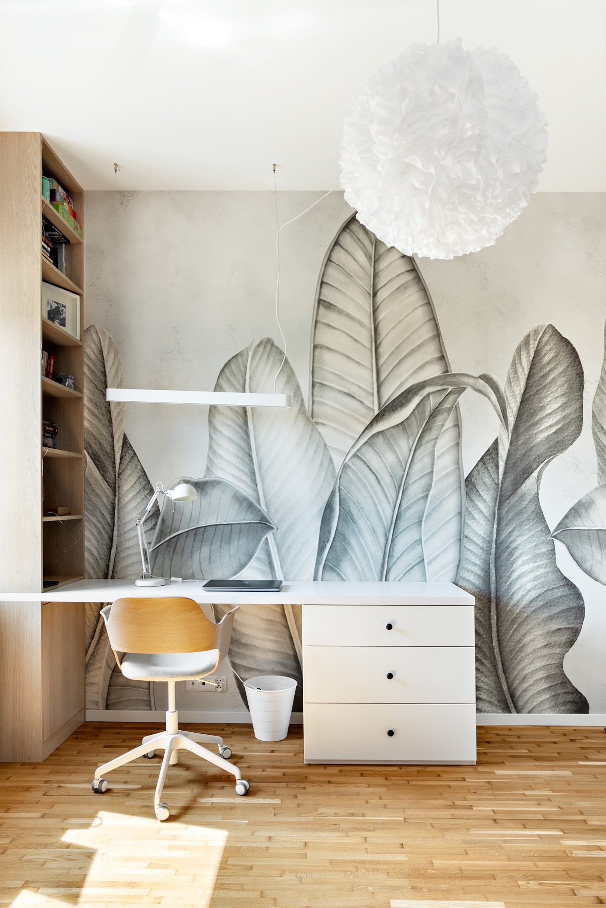 Jasne biuro z drewnianą podłogą, białym biurkiem z szufladami, krzesłem na kółkach i tapetą w liście. Na biurku lampka i laptop, obok kosz na śmieci.