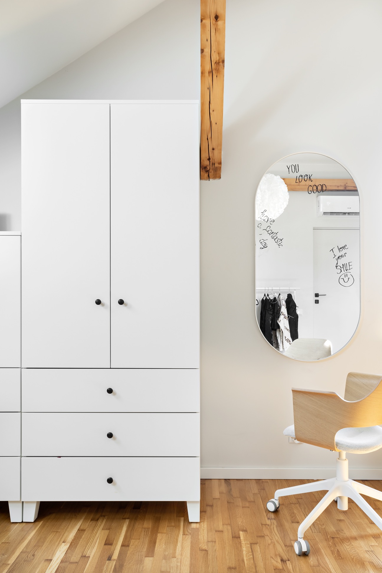 Nowoczesny pokój z białą szafą, owalnym lustrem z napisem 'You look good' i drewnianym krzesłem na kółkach, poddasze z drewnianą belką, parkiet na podłodze.